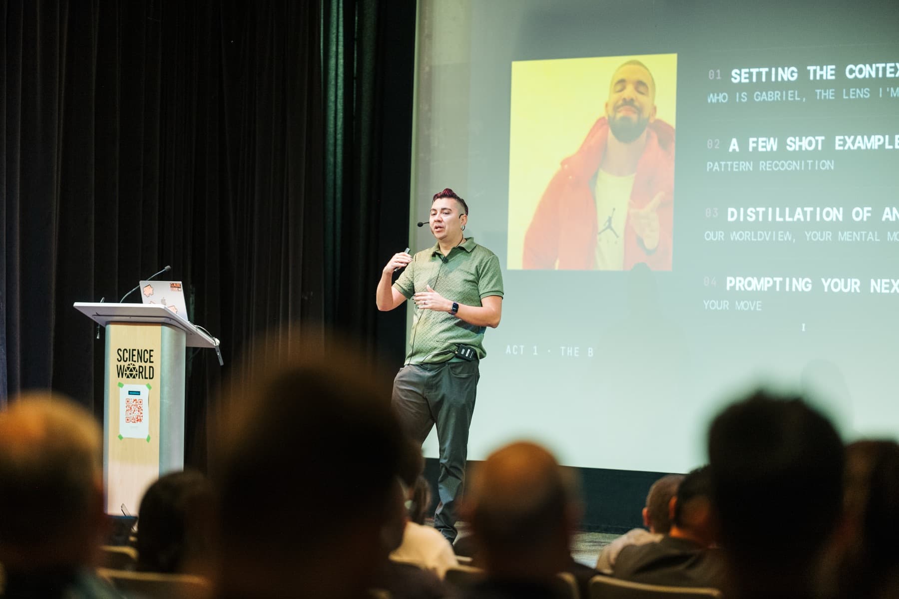 Gabriel Velazquez speaking with a handheld microphone on stage, audience silhouettes in the foreground