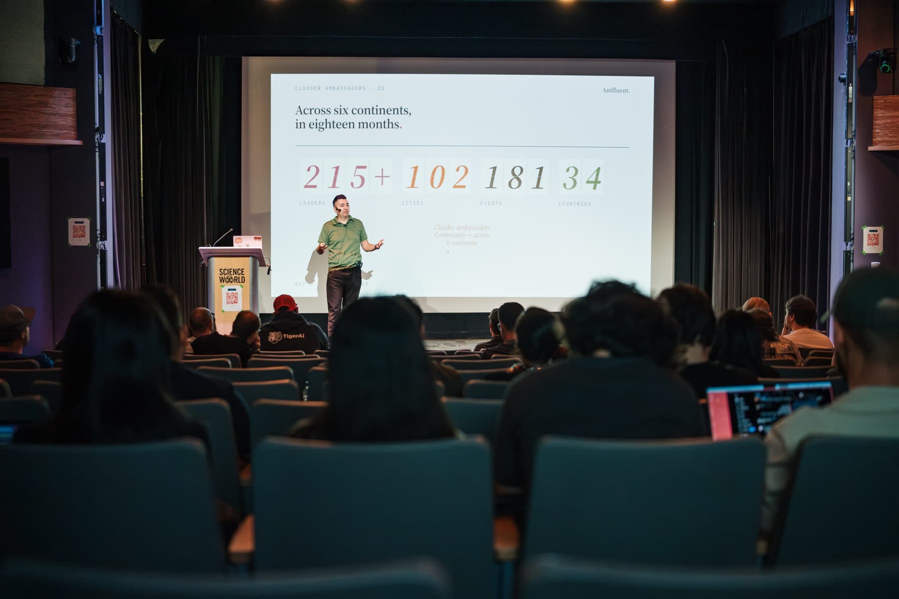 Wide view of the Cloud Summit theatre with Gabriel Velazquez on stage beneath a slide reading Across six continents, in eighteen months