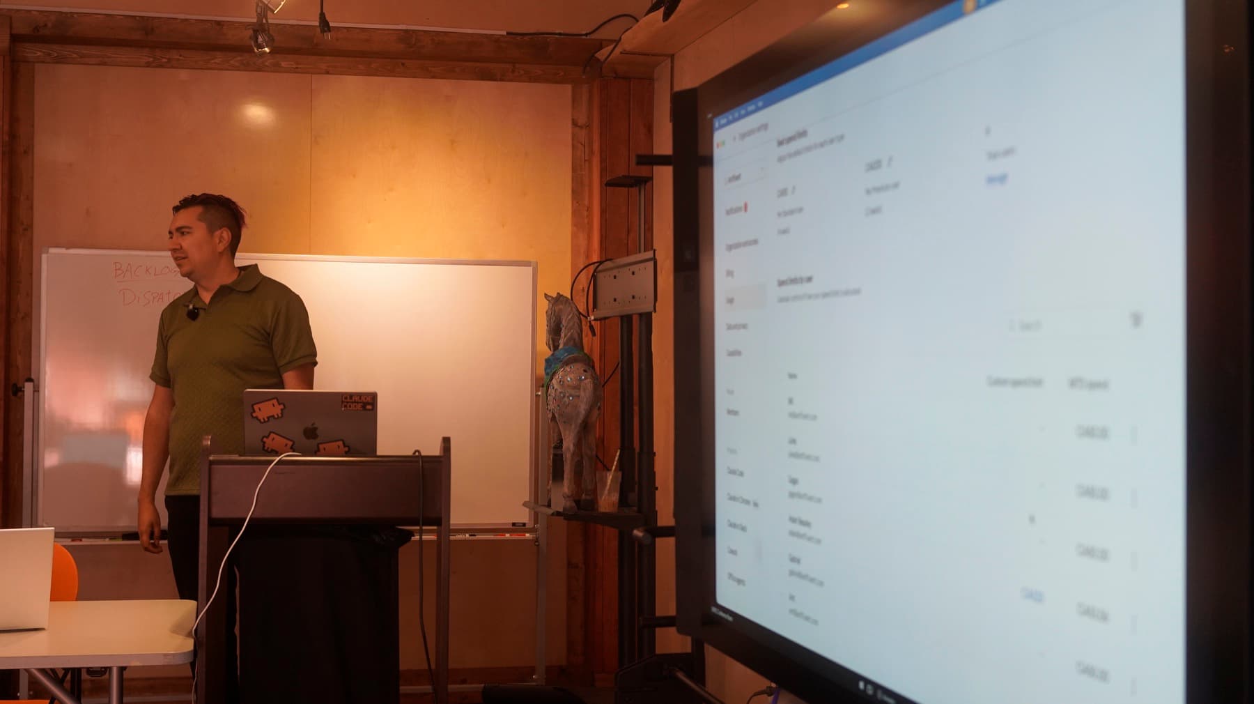 Instructor at a lectern beside a whiteboard, presenting to a large display